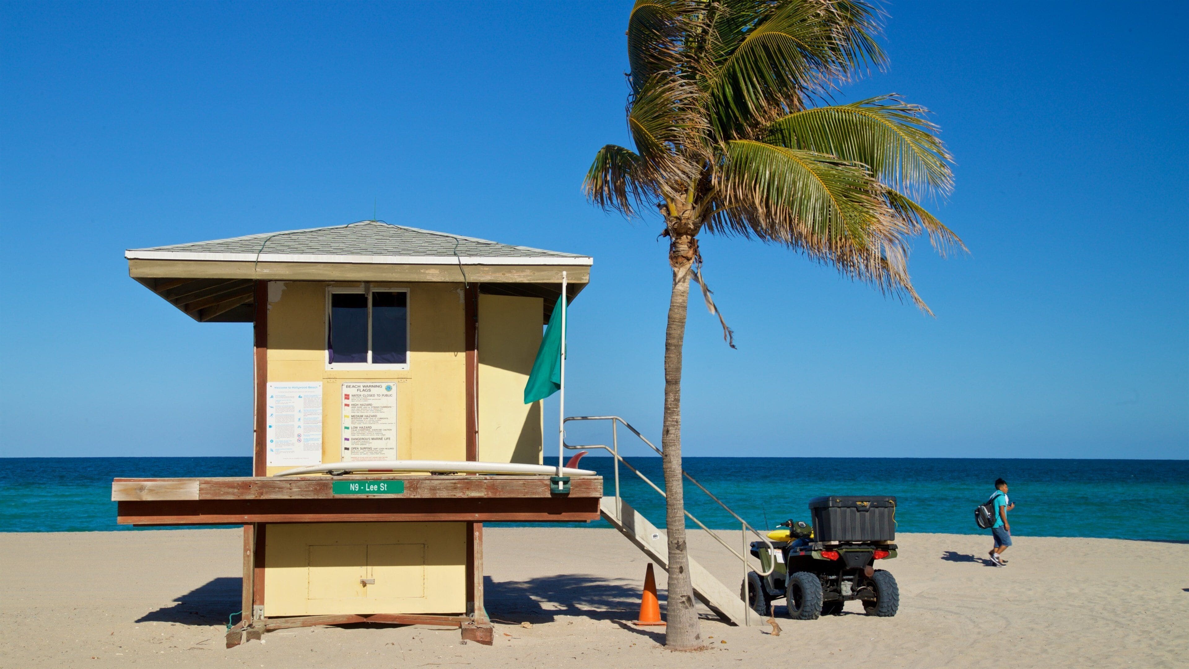 A lifeguard tower on a beach with a palm tree and a person walking by.