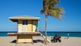 A lifeguard tower on a beach with a palm tree and a person walking by.
