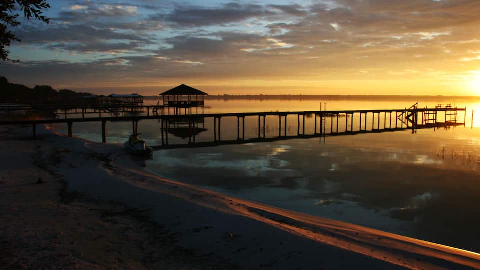 A pier extending into calm waters at sunset with a small shelter and a boat near the shore.