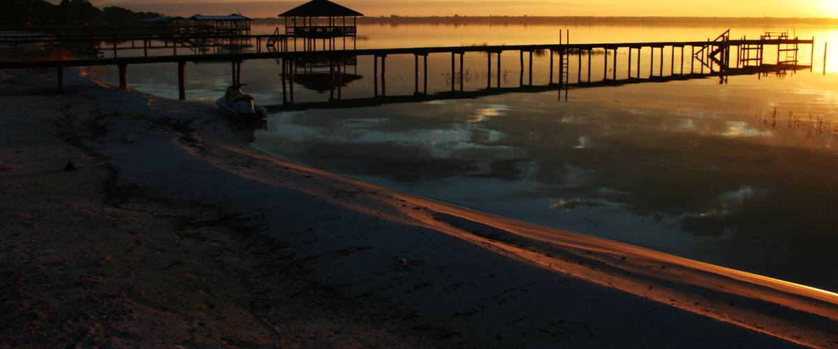 A pier extending into calm waters at sunset with a small shelter and a boat near the shore.