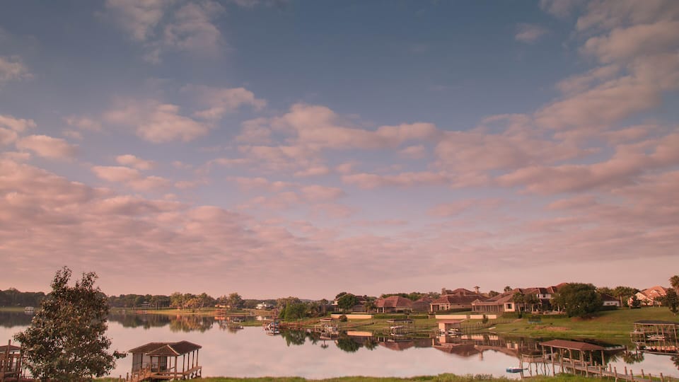 A calm lake with a wooden dock, surrounded by houses and trees under a partly cloudy sky.