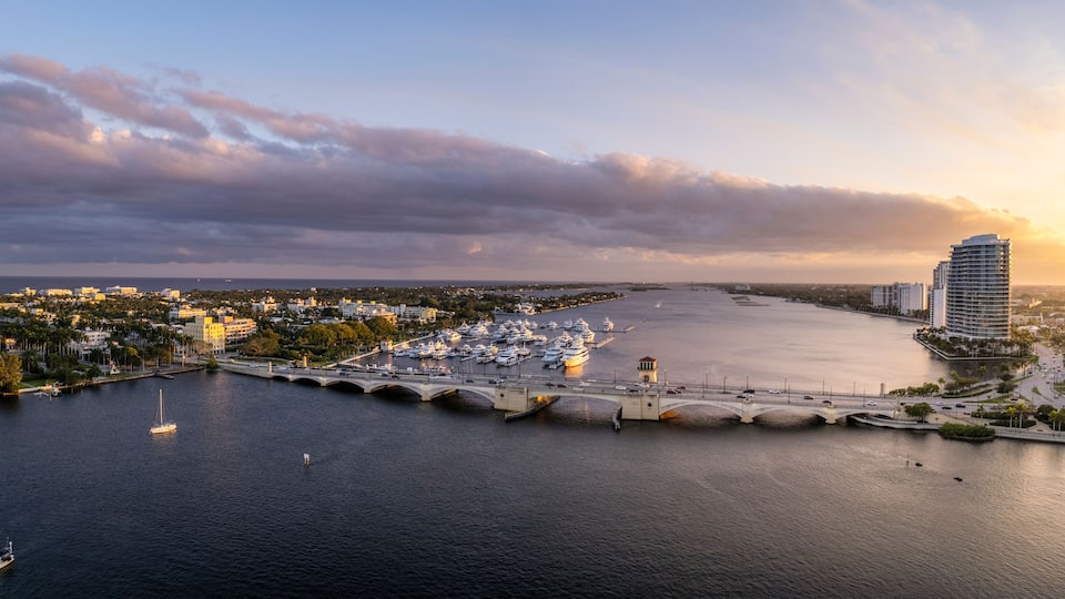 A marina with boats, a bridge, and a cityscape with buildings and a sunset.