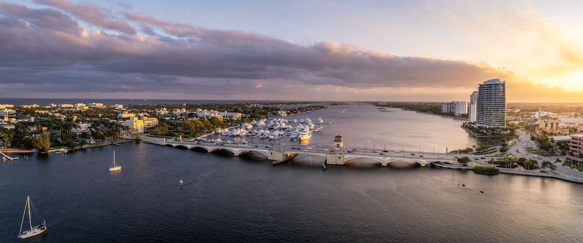 A marina with boats, a bridge, and a cityscape with buildings and a sunset.