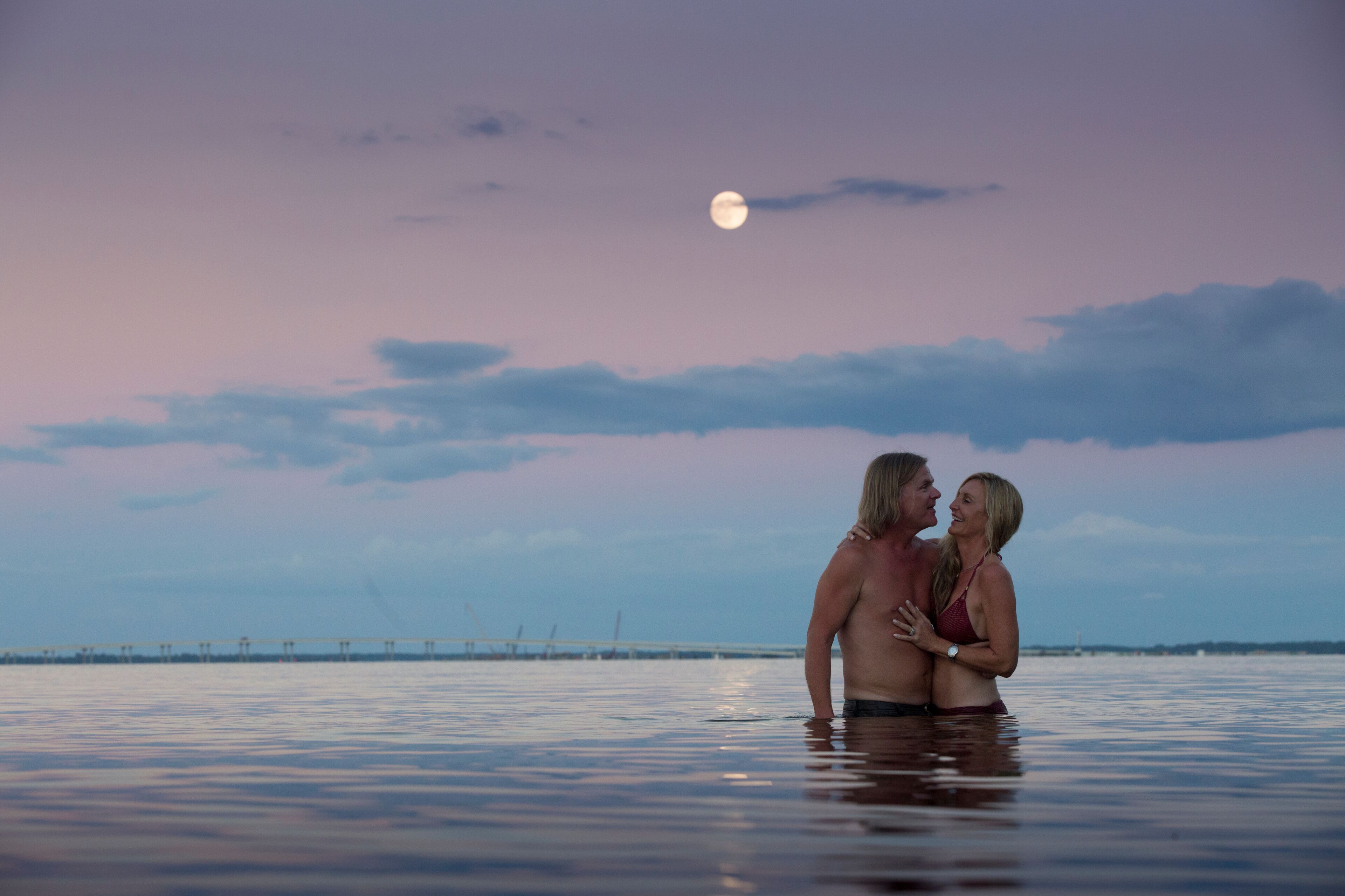 A couple in a body of water at sunset with a bridge in the background.