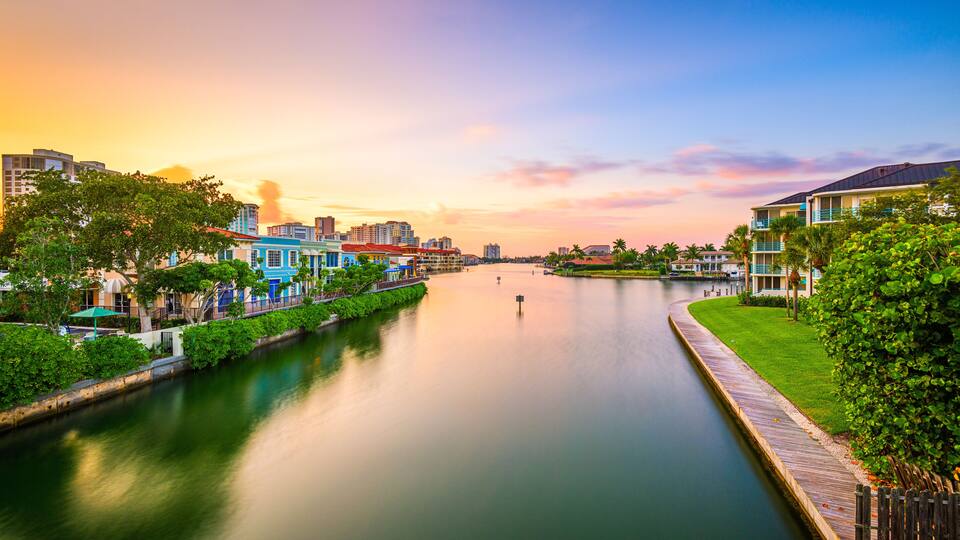 A river with a wooden walkway on the right, surrounded by greenery and buildings with colorful facades.