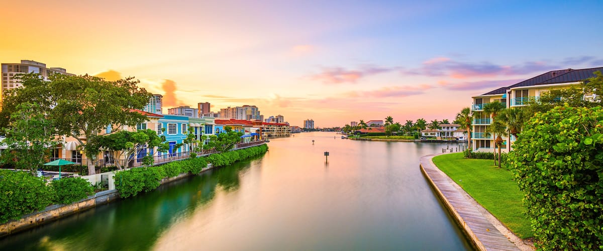 A river with a wooden walkway on the right, surrounded by greenery and buildings with colorful facades.