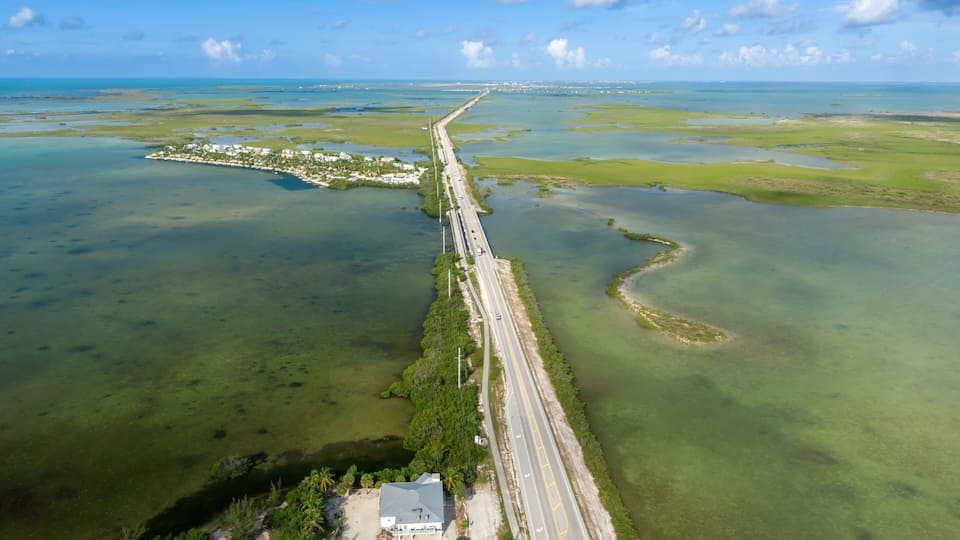 An aerial view of a coastal road with a bridge, surrounded by water and greenery.