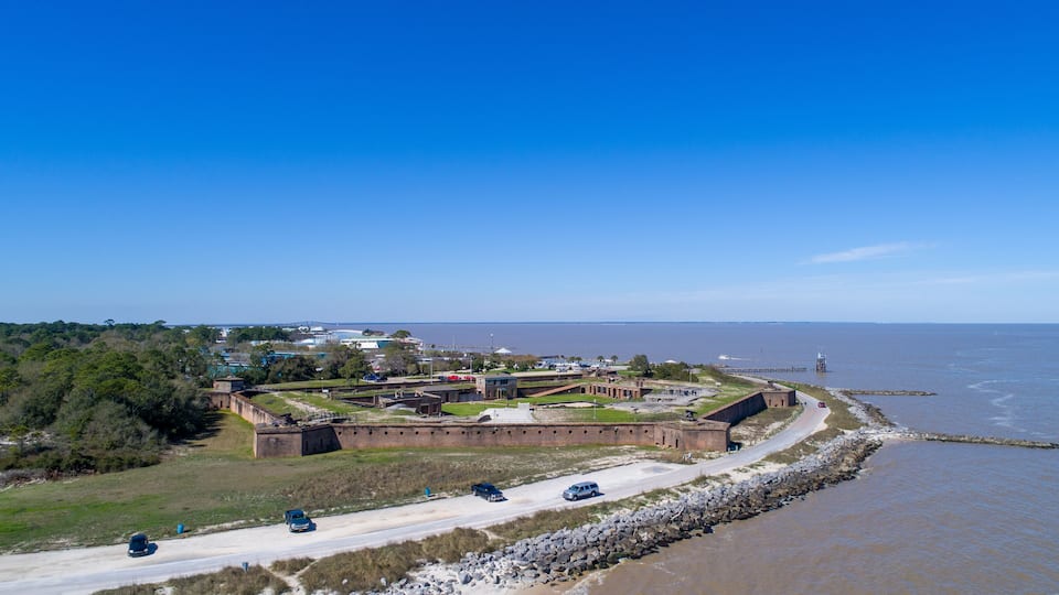 A coastal fortification with a road leading to it, surrounded by trees and a body of water.
