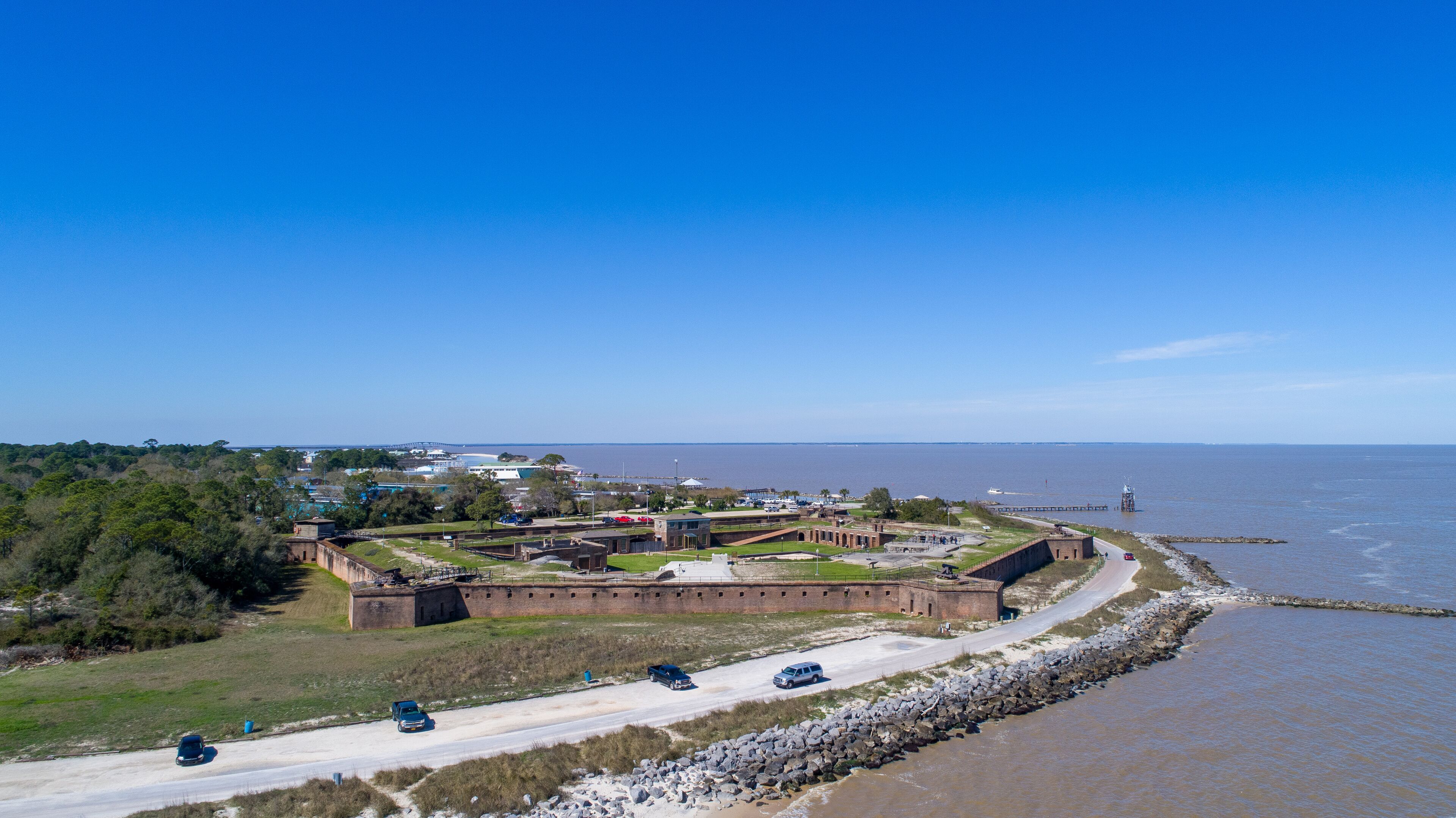 A coastal fortification with a road leading to it, surrounded by trees and a body of water.