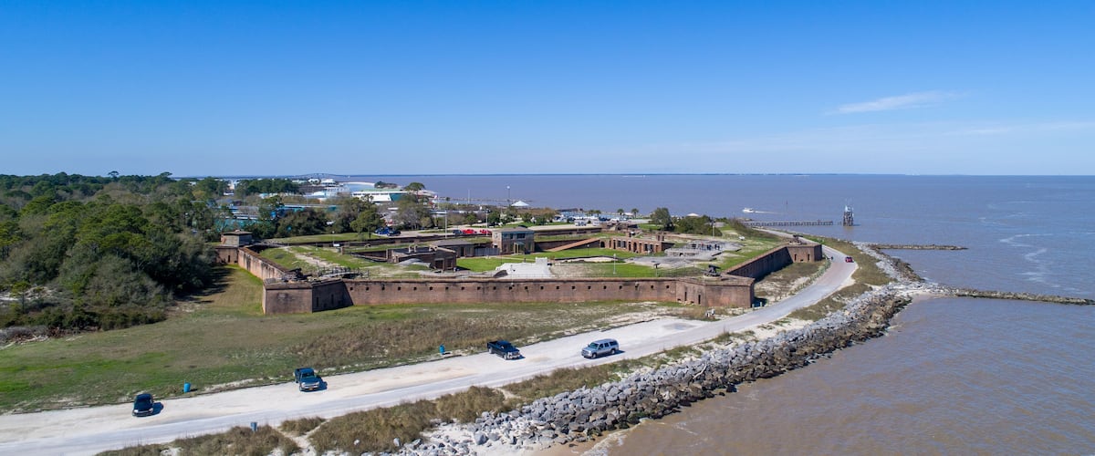 A coastal fortification with a road leading to it, surrounded by trees and a body of water.