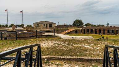 A historical fort with brick buildings, a flagpole with flags, and a coastal view.
