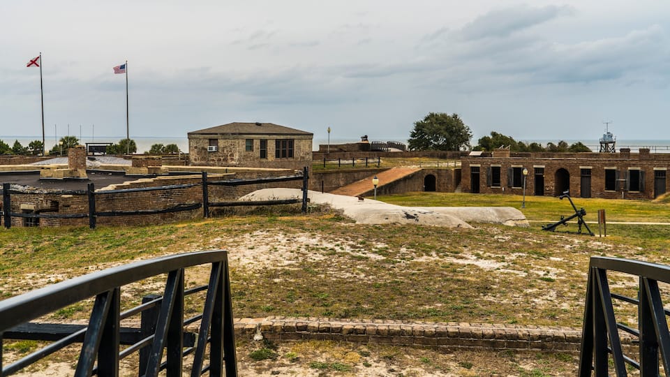 A historical fort with brick buildings, a flagpole with flags, and a coastal view.