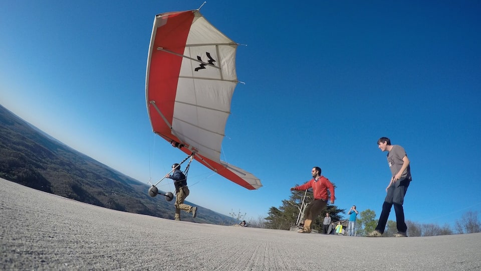 A paraglider preparing to take off with a red and white sail, a mountainous landscape in the background, and a group of people watching on the ground.