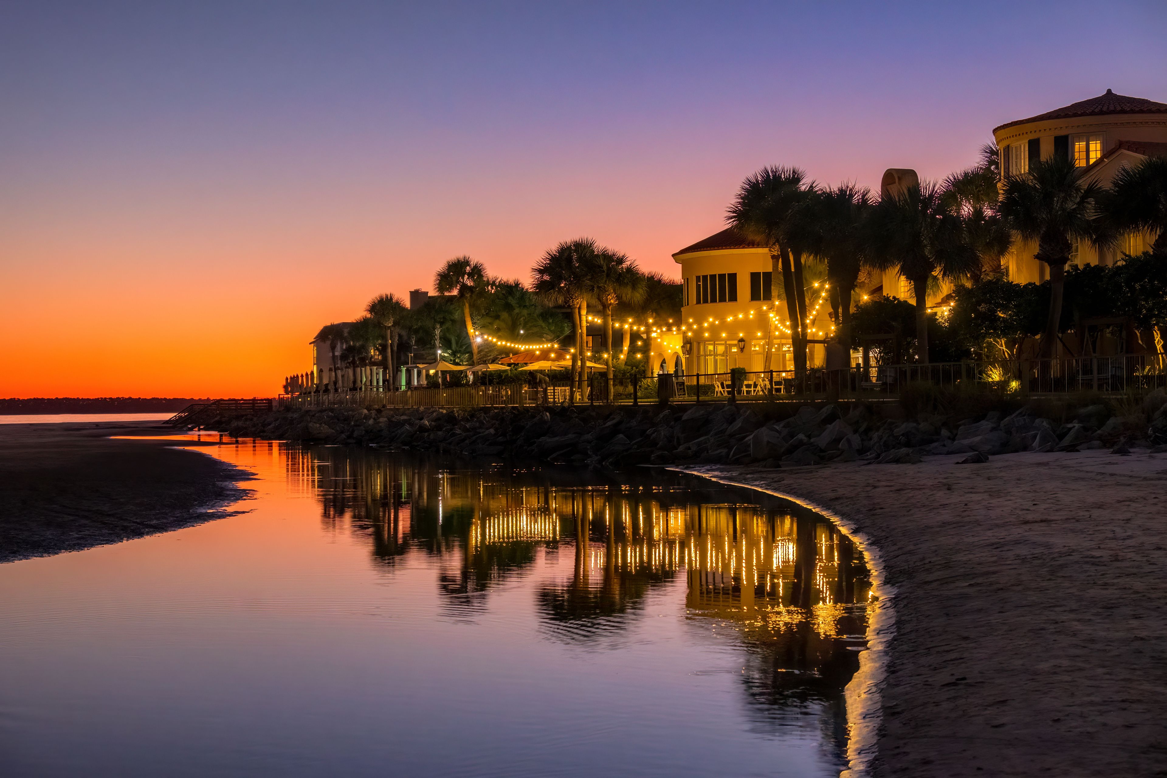 A sunset over a beach with a reflection of lights on the water, palm trees, and a building with string lights.