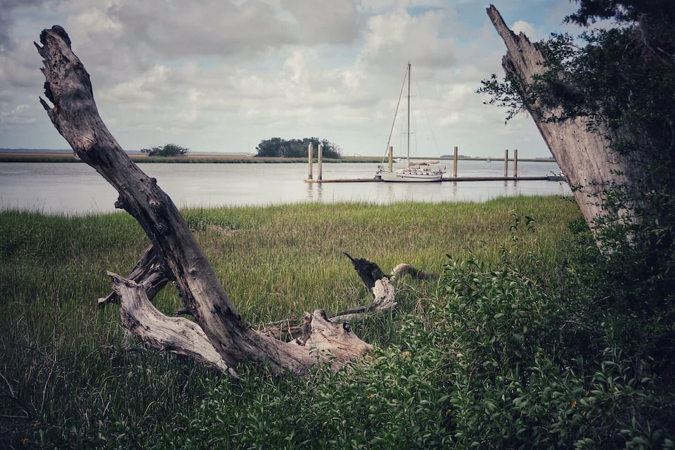 A calm lake with a sailboat docked at a wooden pier, surrounded by grassy banks and a fallen tree trunk.