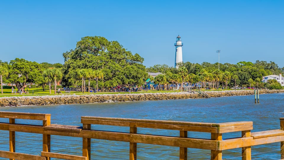 A waterfront park with a lighthouse, trees, and a wooden walkway.