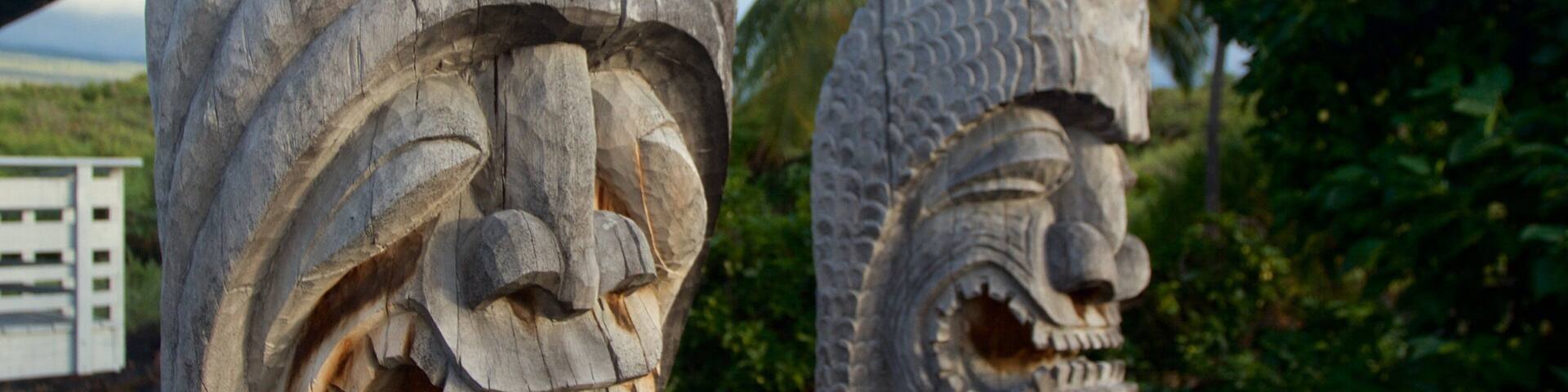Two stone tiki head statues with intricate carvings, set against a backdrop of greenery and a distant view of a body of water.