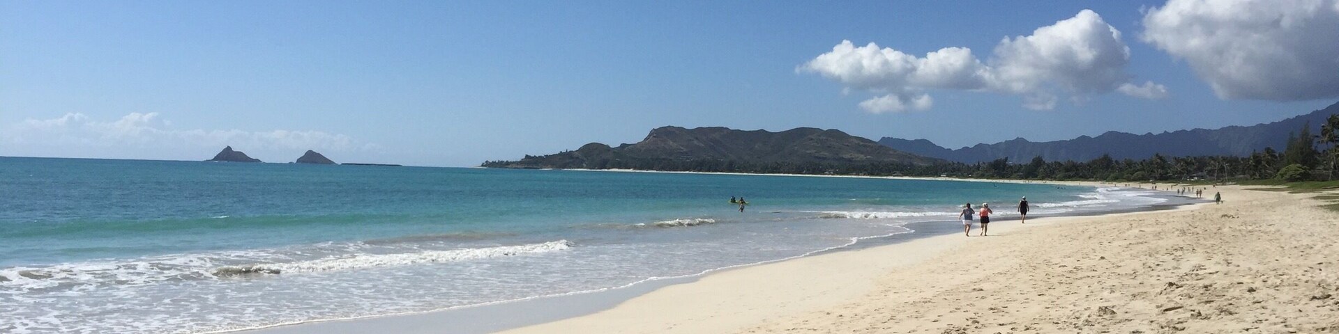 Praia arenosa com pegadas, águas turquesa calmas e montanhas ao longe sob um céu claro.
