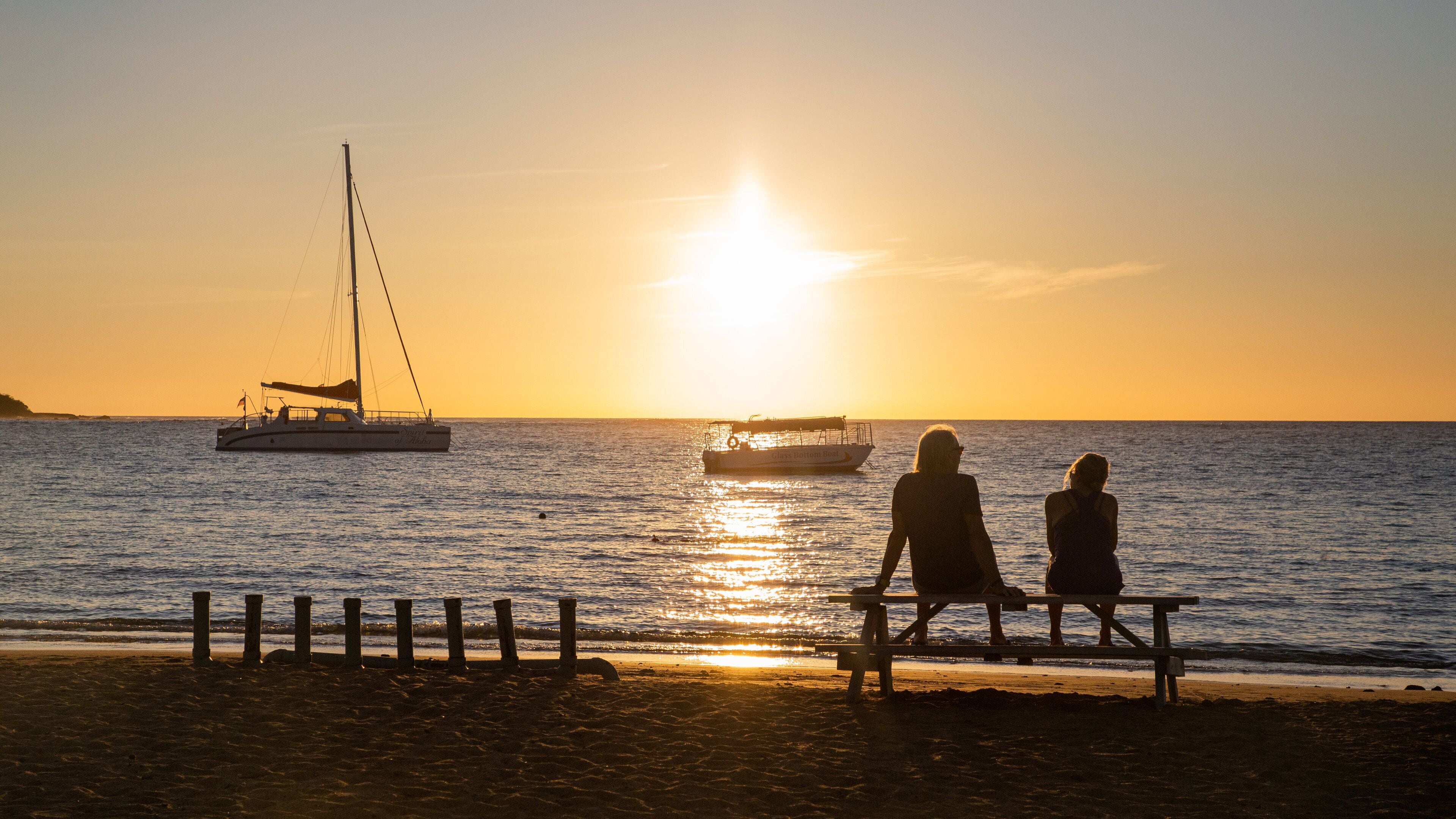 A sunset over the ocean with a sailboat and a smaller boat, two people sitting on a bench facing the water.