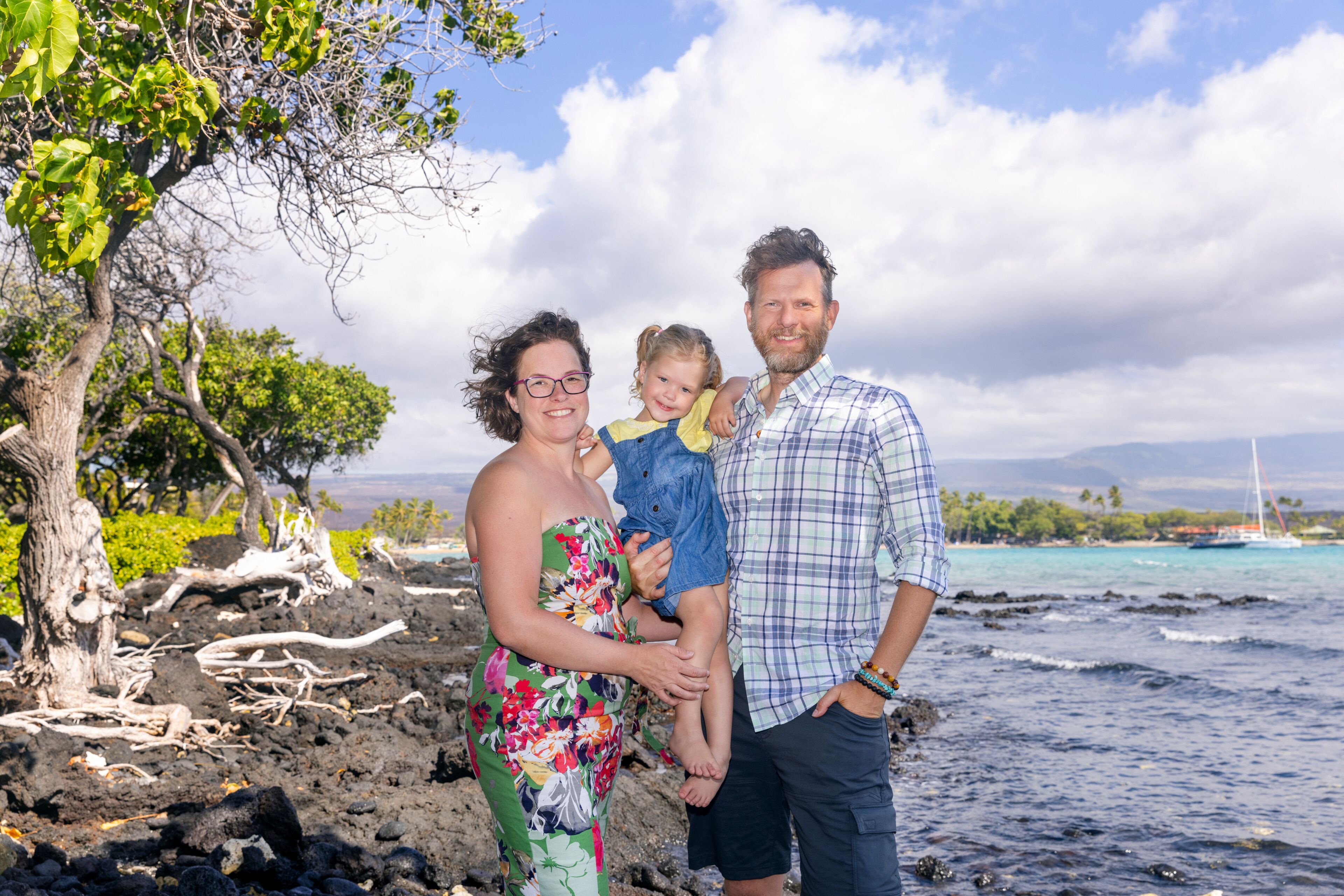 A family by a rocky shoreline with a clear sky and a sailboat in the distance.