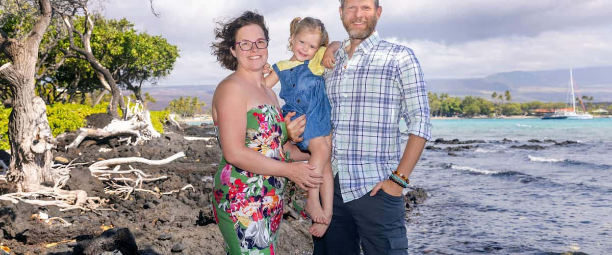 A family by a rocky shoreline with a clear sky and a sailboat in the distance.