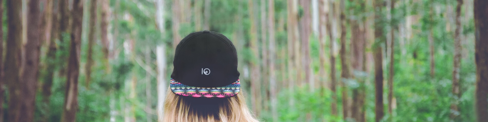 Une personne aux longs cheveux blonds, portant une casquette noire et un haut rouge, se tient dans une forêt où poussent de grands arbres.