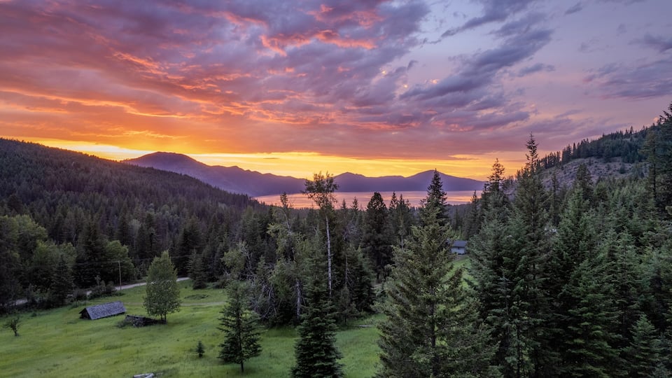 A mountainous landscape with a forested valley, a small cabin, and a lake at sunset.