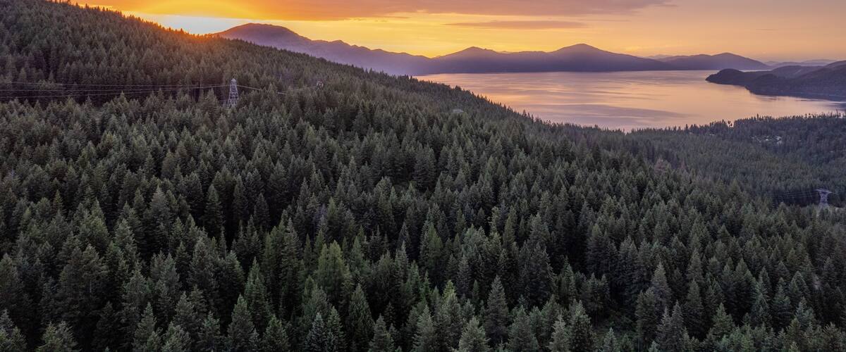 Tiheä, metsäinen vuoristo, tyyni järvi ja kauempana horisontissa kohoavat vuoret auringonlaskun värittämässä taivaassa.
