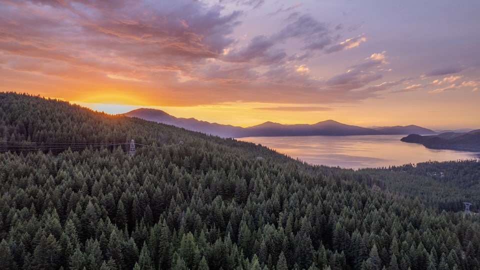 A dense forested mountain range, a calm lake, and distant mountains under a sunset sky.