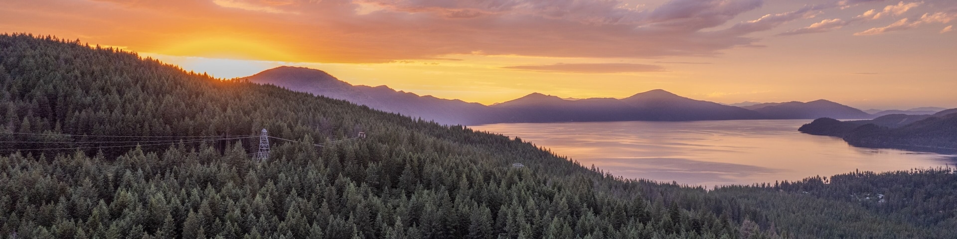A dense forested mountain range, a calm lake, and distant mountains under a sunset sky.