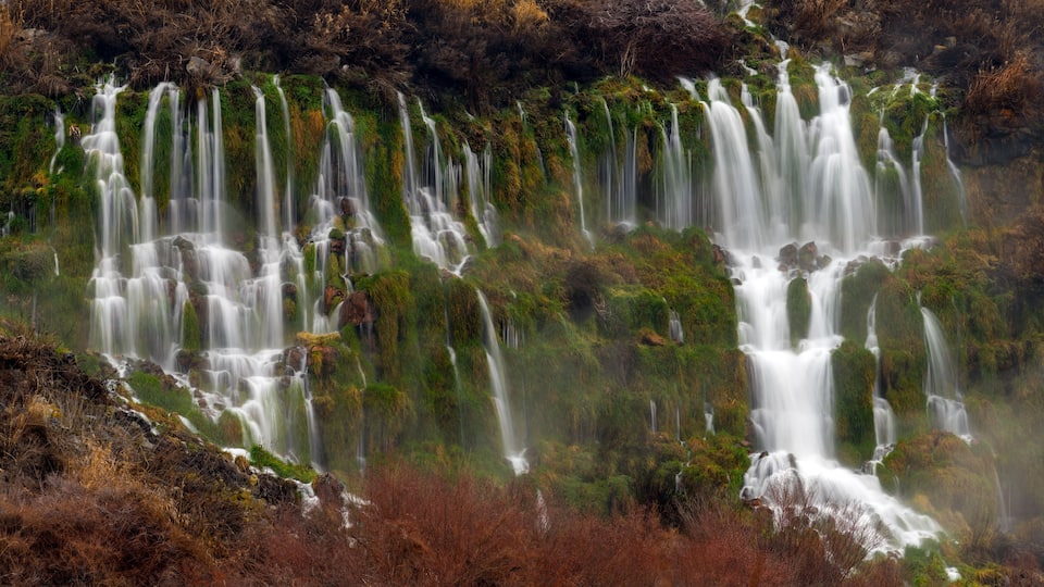 A multi-tiered waterfall with moss-covered rocks and surrounding vegetation.