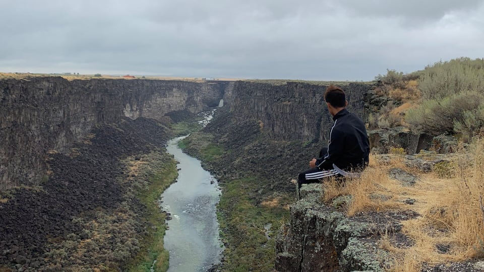A person sitting on a cliff overlooking a deep canyon with a river flowing through it.