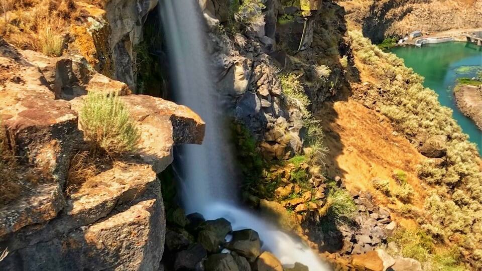 A waterfall cascading down a rocky cliff into a turquoise pool surrounded by dry, grassy terrain.