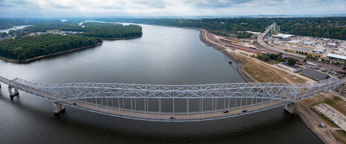 A large suspension bridge over a river, surrounded by greenery and industrial areas.
