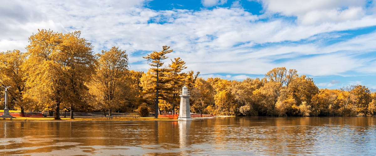 Een rustig meer met herfstbomen, de reflectie in het water, een vuurtoren in het midden en een gedeeltelijk bewolkte lucht.