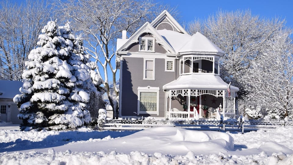 A snow-covered house with a porch and a snow-covered tree in front.