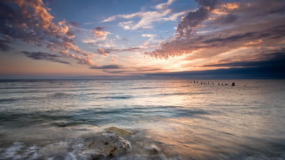 A sunset over a calm sea with a rocky foreground and a row of poles in the distance.
