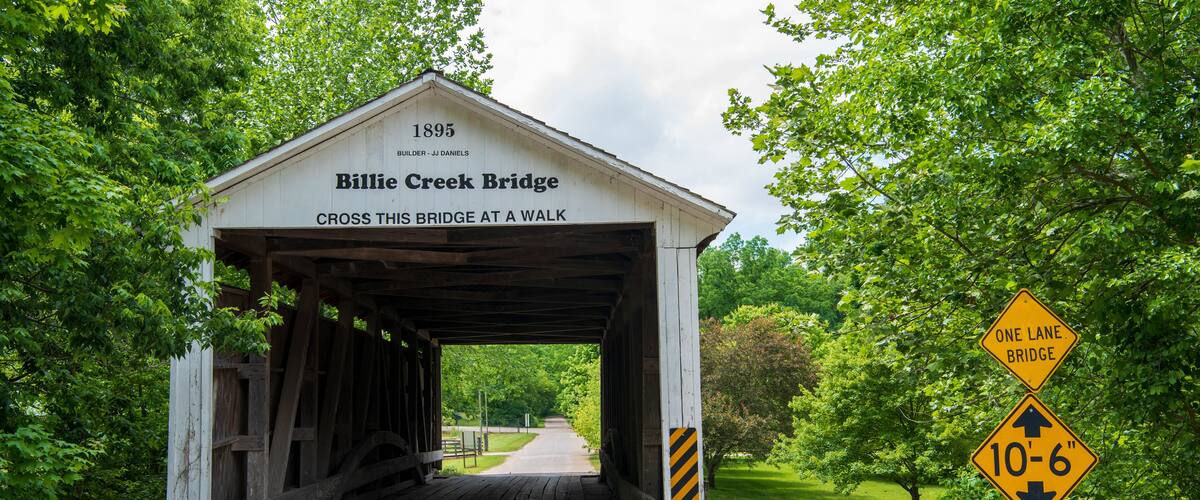 A covered bridge with a sign reading "Billie Creek Bridge 1895 BUILDER - JJ DANIELS CROSS THIS BRIDGE AT A WALK."