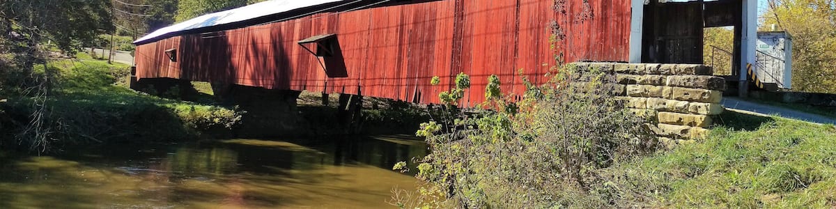 Eine rote gedeckte Brücke über einem Fluss, mit einer Steinmauer und umliegender Vegetation.