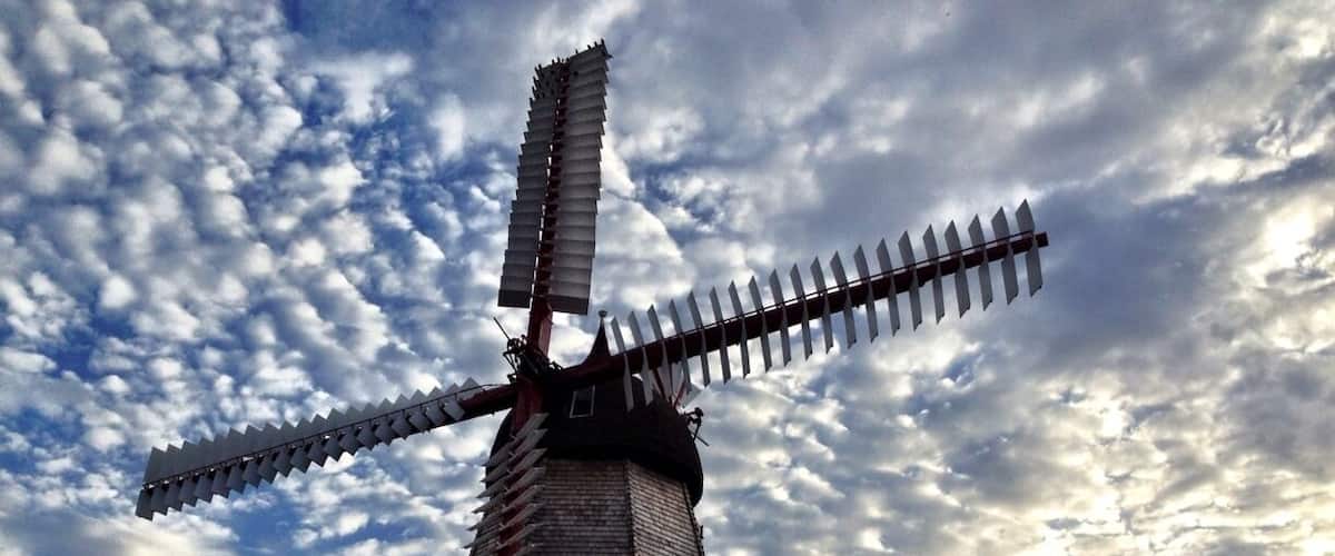 A traditional windmill with a wooden structure and a large sail, set against a cloudy sky.