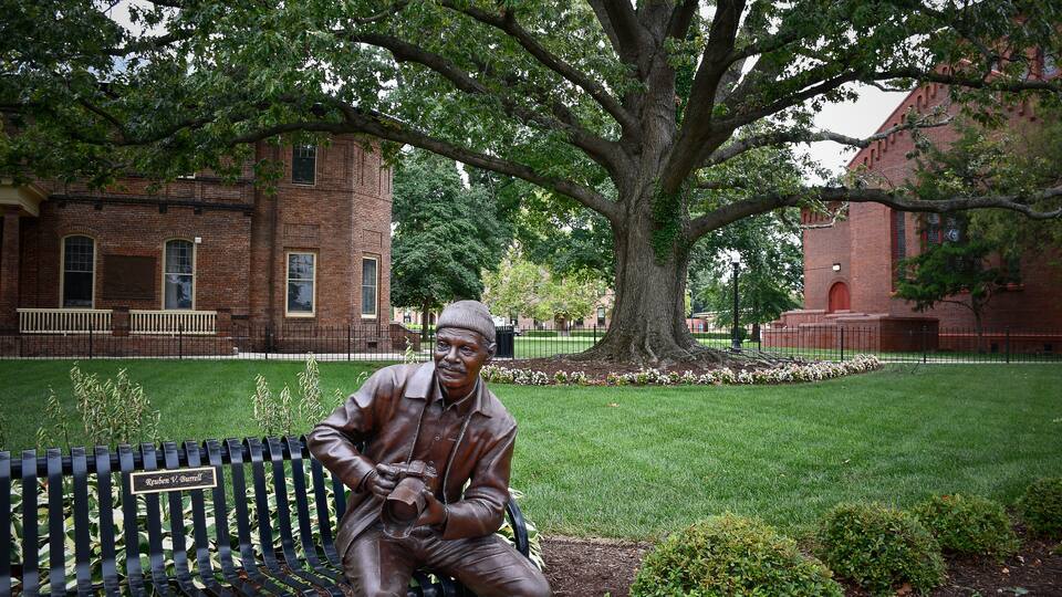 A bronze statue of a man sitting on a bench with a nameplate, a large tree, and a brick building in the background.