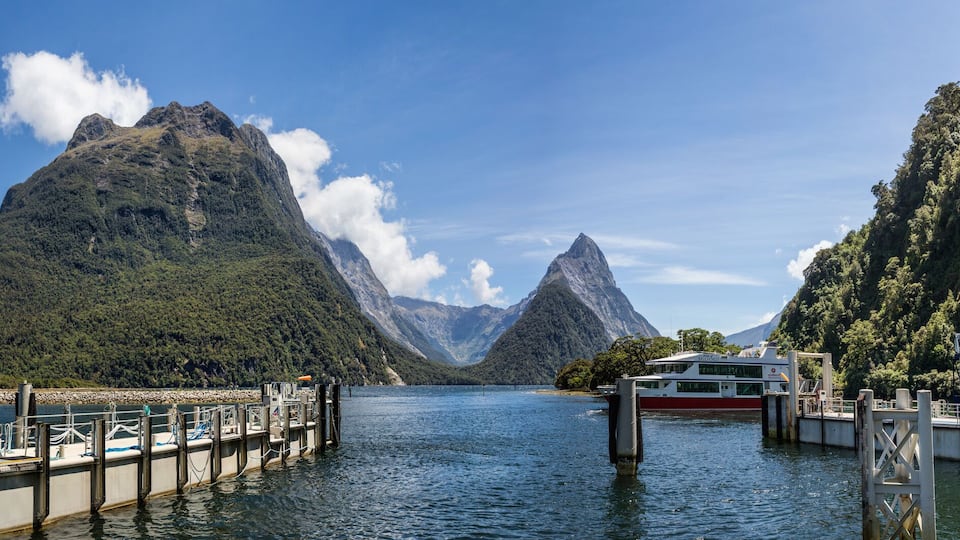 A river with boats docked at a pier, surrounded by steep, forested mountains.