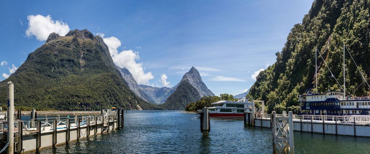 A river with boats docked at a pier, surrounded by steep, forested mountains.