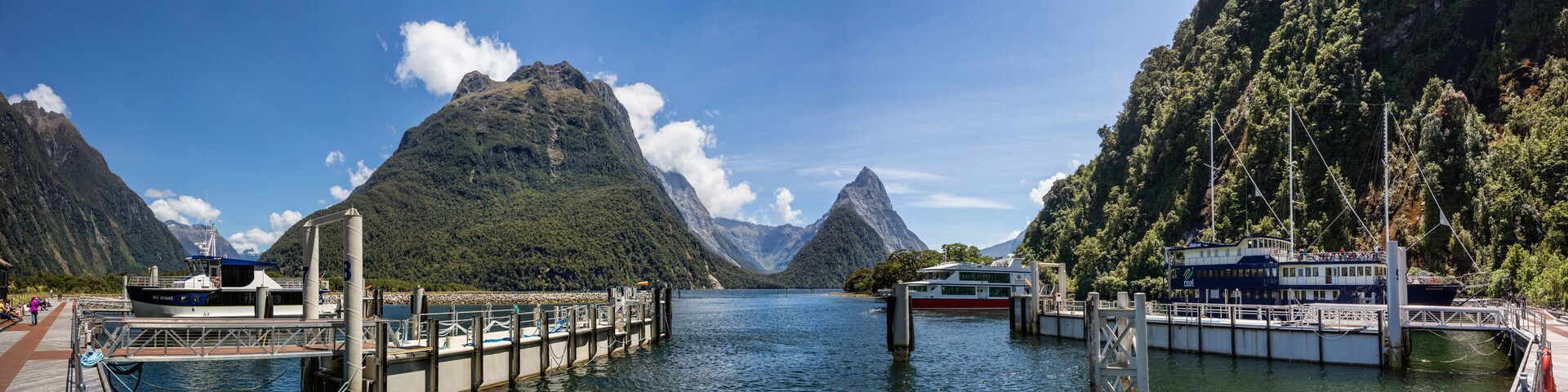 A river with boats docked at a pier, surrounded by steep, forested mountains.
