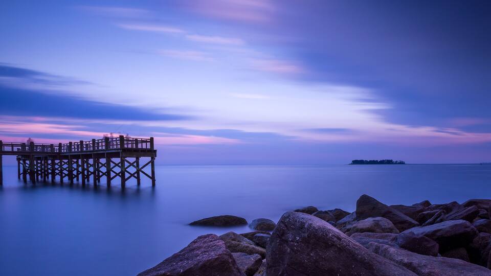 A pier extending into calm waters with a rocky foreground and a distant island.