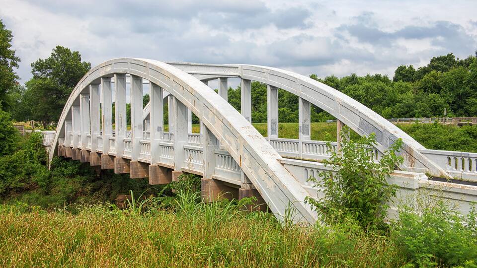A white arched bridge with railings, surrounded by greenery and trees.