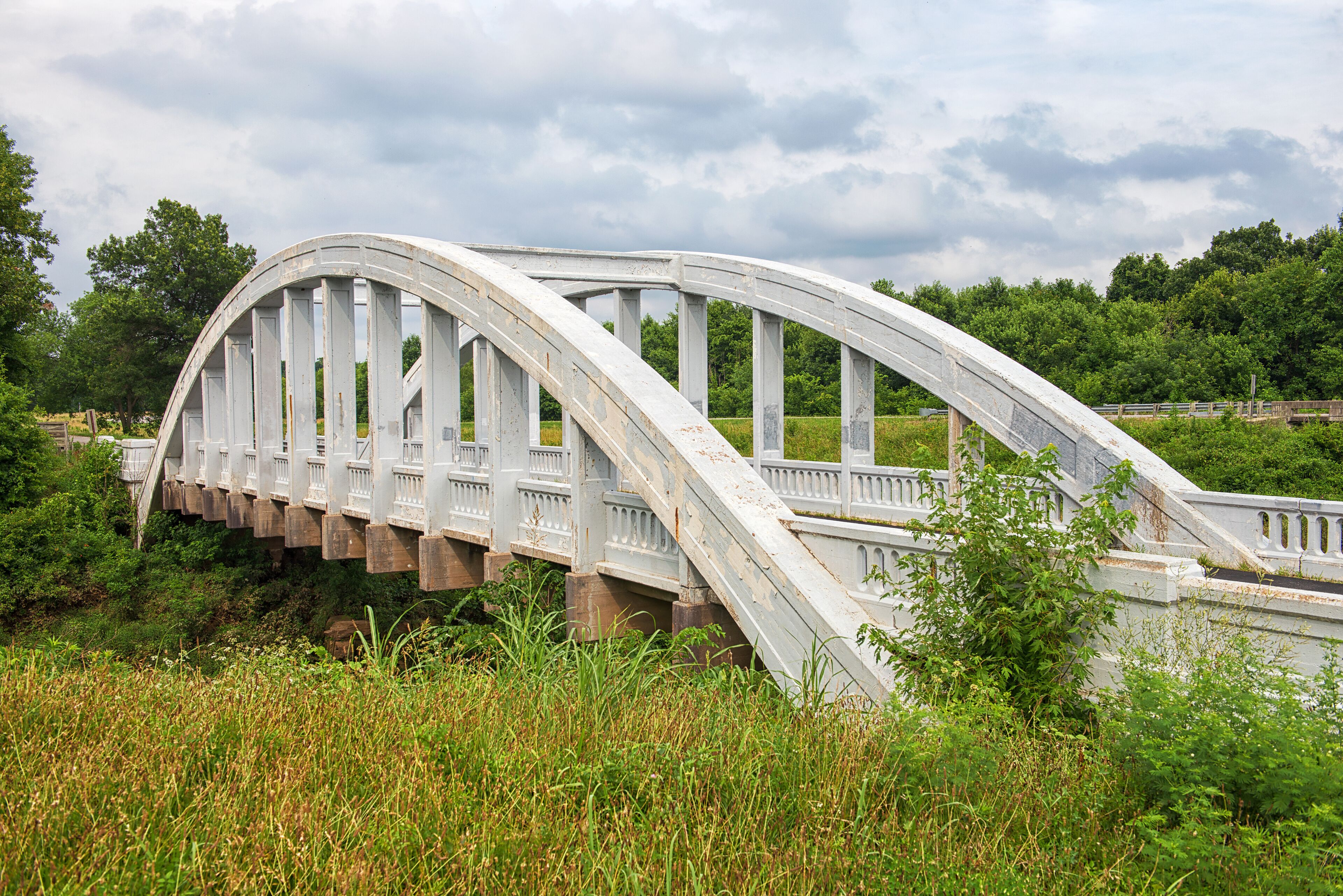 A white arched bridge with railings, surrounded by greenery and trees.