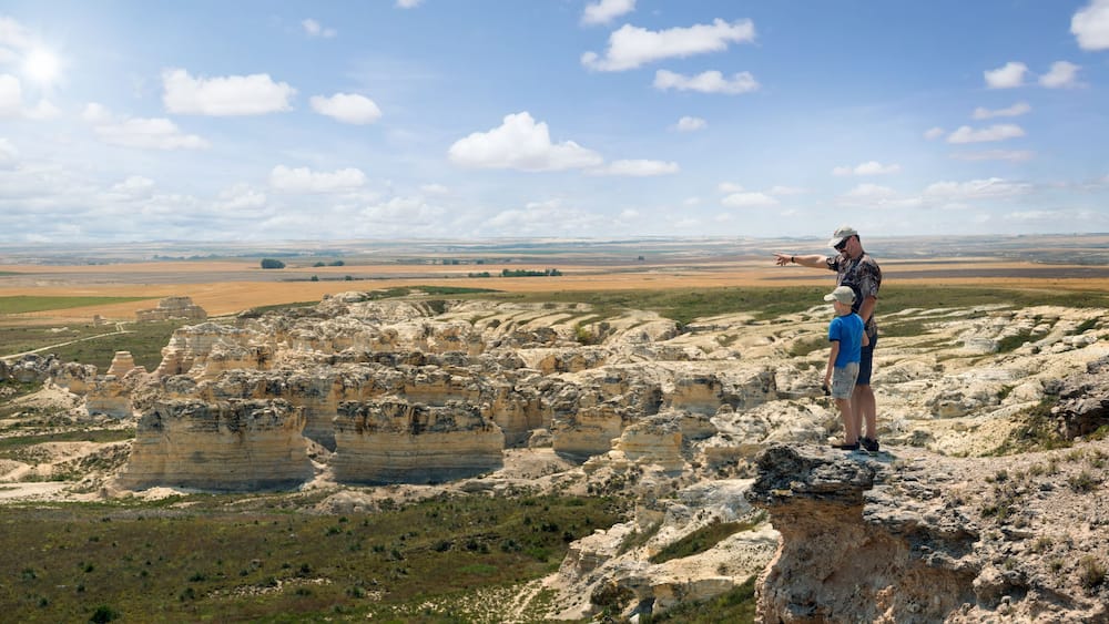 A man and a child standing on a rocky outcrop overlooking a vast landscape with layered rock formations and distant fields.
