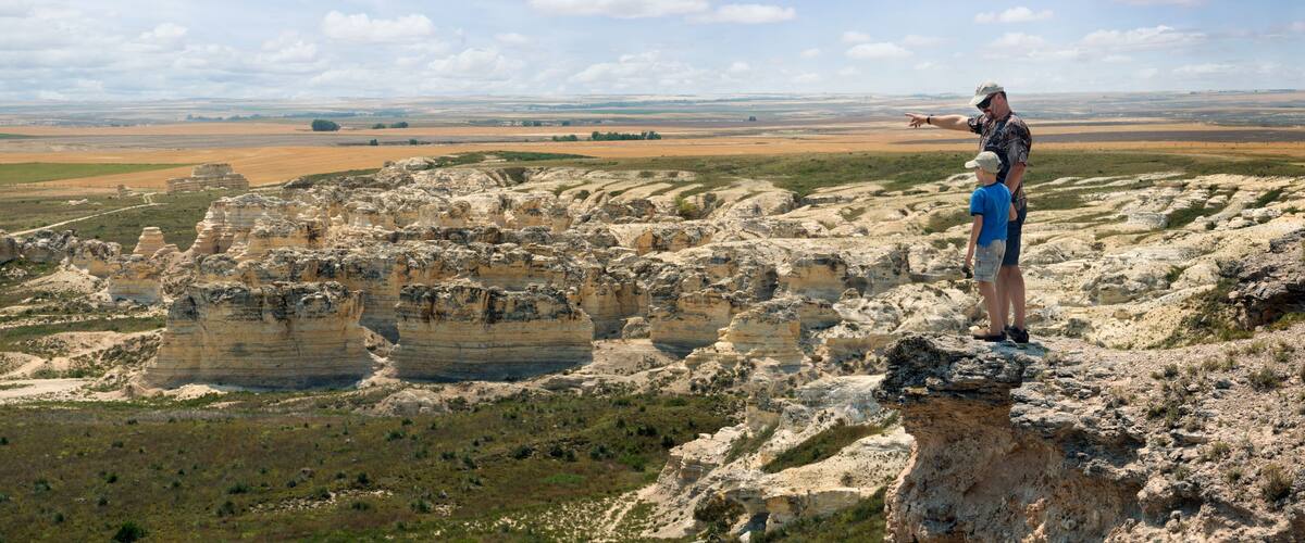 A man and a child standing on a rocky outcrop overlooking a vast landscape with layered rock formations and distant fields.
