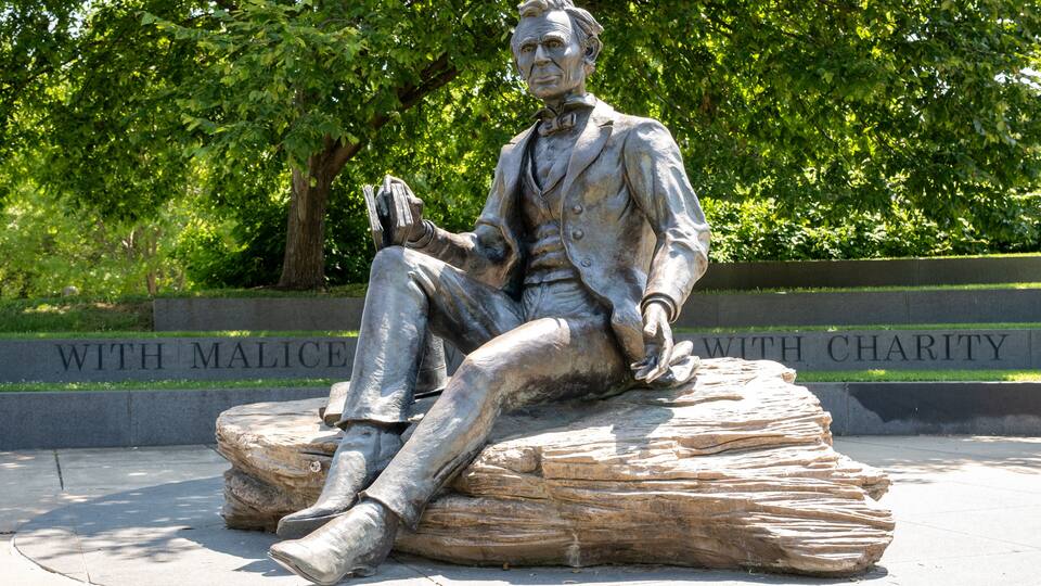 A statue of a seated man in a suit with a book, set on a rock with inscriptions in the background.